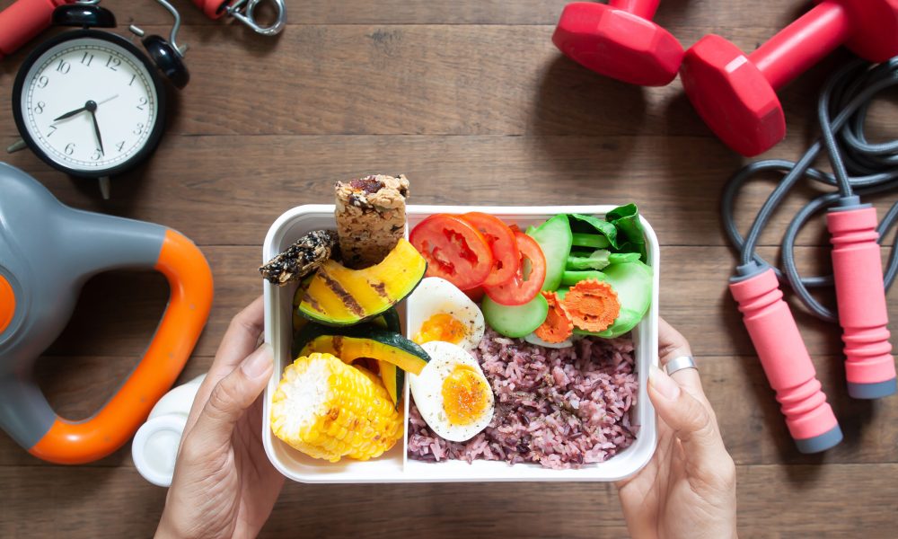 Woman's hands holding lunch box with rice berry, boiled eggs, sweetcorn, pumpkin, tomatoes and cereal bars, Top view with sport and fitness equipments on wooden background
